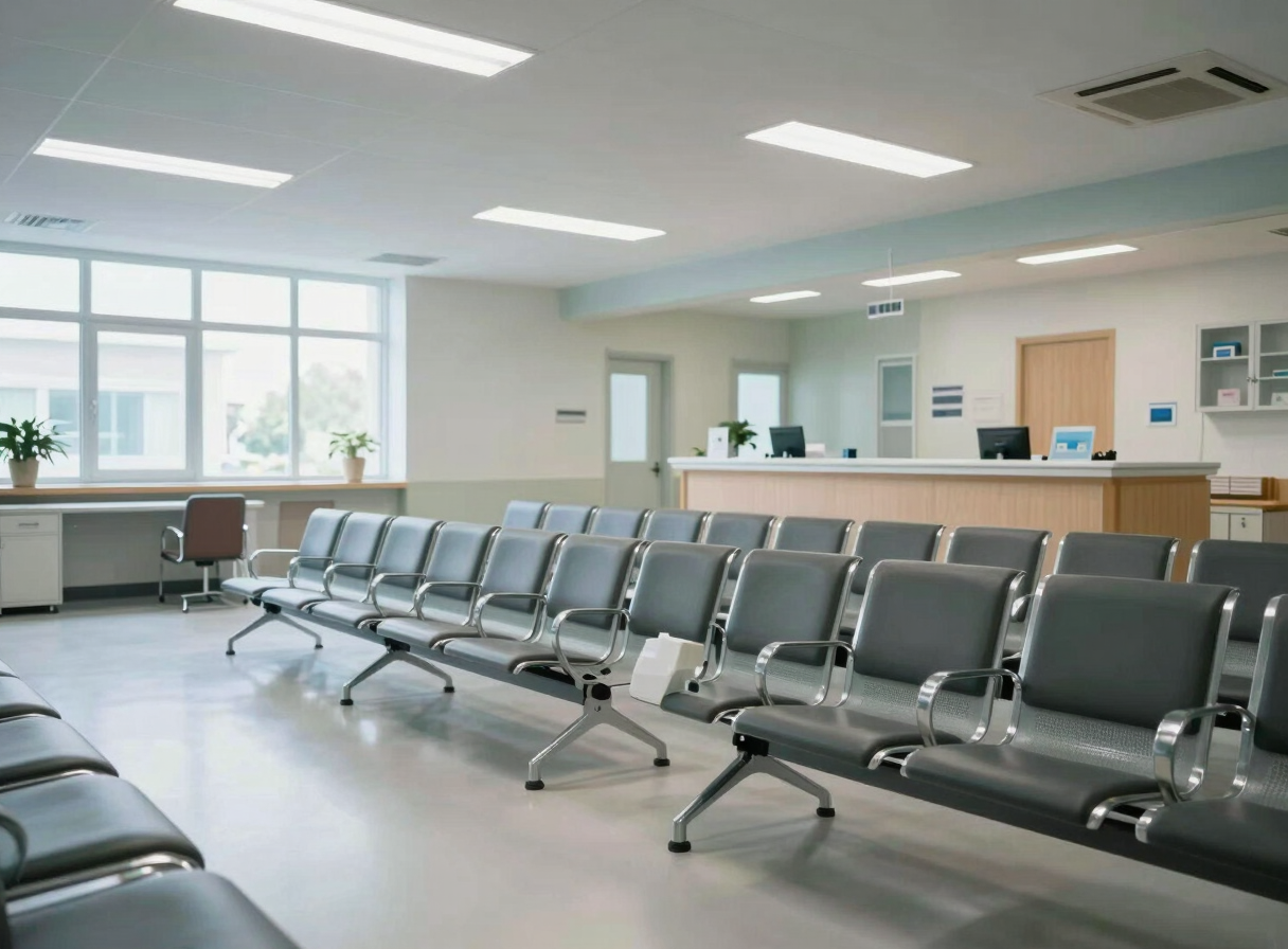 Wide environmental shot of a clean clinical waiting area interior, rows of padded chairs, reception desk visible in background, north-facing window light, cool fluorescent overhead, no people present, clinical modern decor