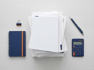 Clean corporate office flatlay with neatly stacked reams of white paper, a blue pen, and a small calculator, bright high-key studio lighting, organized layout, crisp navy and orange accents, sharp focus.