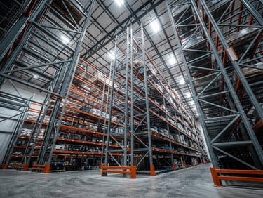 Low-angle wide shot looking up at massive industrial warehouse racking structures, heavy-duty steel beams, cool steel tones, high-contrast overhead lighting.