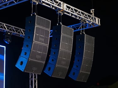 Professional line array speakers hanging from a stage truss, dramatic dark background with subtle blue neon highlights, sharp details.