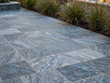 Completed hardscape patio in natural daylight — large-format bluestone pavers with tight sand-set joints, low retaining wall edge, coastal grasses planted along border, no people