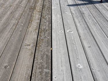 Before-and-after pressure washing split — left half showing weathered gray deck boards with mildew staining, right half showing the same boards clean and bright in afternoon daylight, no people