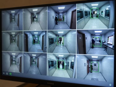 Extreme close-up of a CCTV monitor bank in a hotel security room, screens showing multiple live feed tiles of corridors and kitchen areas with green bounding-box overlays on staff positions, ambient blue-grey monitor glow, no faces identifiable