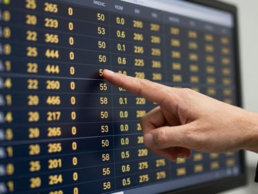 Close-up of a professional's hand pointing at a screen displaying real-time gold market data, with a subtle reflection of gold jewelry, clean office lighting, deep navy and warm gold tones, sharp focus, 50mm lens.