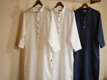 Hanging custom kurtas and ethnic garments made of soft handloom fabrics, casting soft shadows on a warm plaster wall