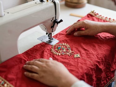 Close-up of hands guiding heavy embroidered crimson fabric through a sewing machine in a sunlit Haveli workshop