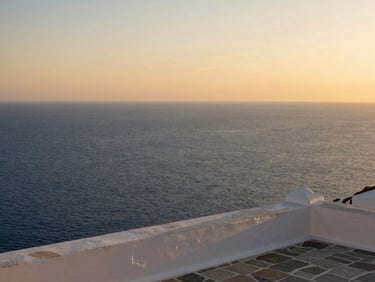 Wide editorial shot of the Aegean Sea at golden hour viewed from a stone terrace on Naxos, deep blue water stretching to the horizon on the left two-thirds, a whitewashed parapet wall in the lower right, warm amber sky, no people, calm and spacious