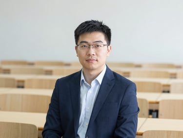 Professional headshot of an East Asian male academic in a modern lecture hall setting, soft background, sharp professional attire