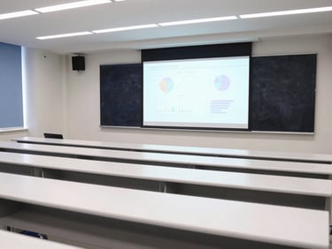 A bright, modern university lecture hall with rows of clean white desks and a wide presentation screen showing scientific data graphs.