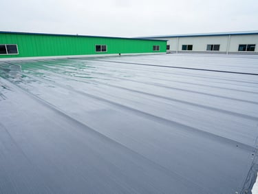 Wide shot of a large industrial warehouse roof with newly applied seamless grey waterproofing membrane, sharp perspective, high-contrast cool tones.