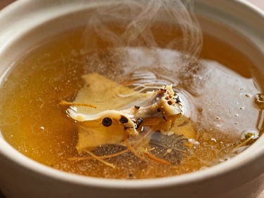 Rich golden bone broth steaming in a ceramic bowl, macro shot showing glistening surface and herbs, dramatic side lighting.