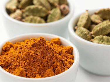 Macro shot of vibrant orange turmeric powder and green cardamom pods in minimalist ceramic cups under natural daylight.