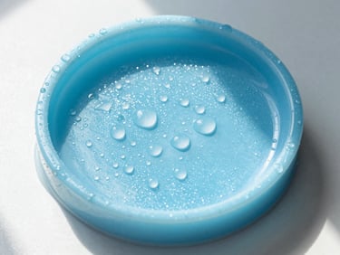 Overhead shot of a translucent sky-blue resin soap dish with water droplets, catching bright morning sun on a white surface