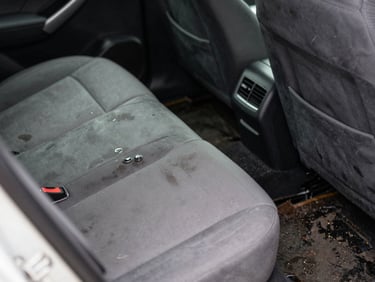 Close-up of a car interior with waterline stains on the seat fabric and mud residue on the floor mat, natural daylight through the window, functional documentary framing