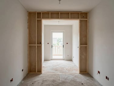 Wide interior shot of a house mid-full-renovation, stripped walls, new stud partitions framed, skip visible through the front door, daylight flooding the empty hallway