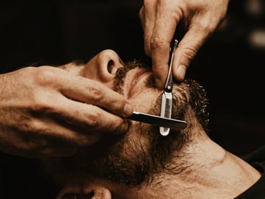 Close-up of a barber's hand meticulously profiling a beard with a straight razor, sharp focus on the blade and skin, warm golden light, deep black background, 35mm.