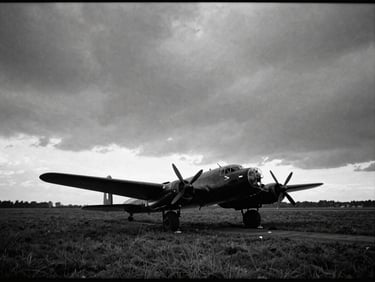 Archival monochrome photograph, vintage film grain, wide atmospheric shot of a grass airfield at dusk, the dark silhouette of a Lancaster bomber resting under a dramatic overcast sky.
