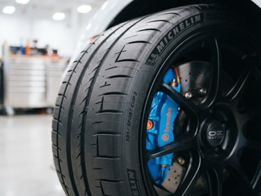 Sharp close-up of a pristine sports car tire tread pattern under bright high-key workshop lighting, vivid blue and orange accents on the brake caliper behind it, 35mm