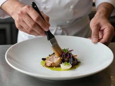 Tight detail of a chef's hands plating a dish on a wide ceramic plate, soft north-facing restaurant kitchen light, sauce brush mid-stroke