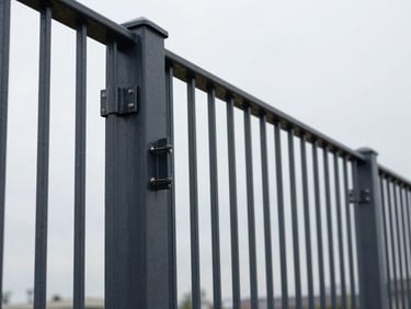 Low-angle shot of a dark powder-coated steel fence section, perfectly aligned posts stretching into the distance, crisp overcast daylight highlighting the texture, heavy industrial latch in foreground, clean lines.