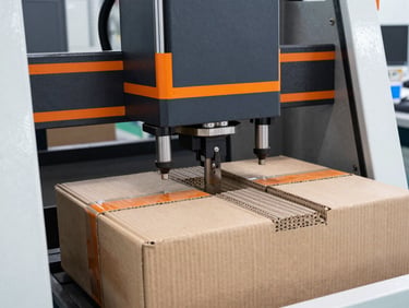 Sharp industrial photography of a heavy hydraulic compression testing machine crushing a corrugated carton, safety-orange accents, bright clean-room lighting.