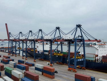 Overhead view of a busy industrial port terminal with multiple cranes loading containers onto a ship, overcast daylight, wide-angle cinematic composition, deep navy and steel tones.