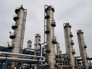 Wide-angle shot of large, interconnected industrial distillation columns against an overcast sky, steel tones, focused on the complex piping of a chemical plant.