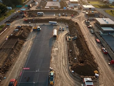 Wide-angle overhead drone frame of an active construction site mid-phase — graded earthworks, foundation grid, and equipment tracks visible from directly above, golden-hour natural light raking low across the site, no people, documentary framing