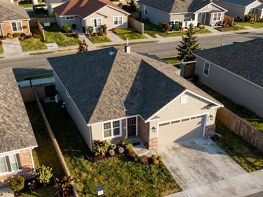 Overhead perspective of a residential property showing roofline, landscaping, driveway, and lot boundary in clear afternoon light, surrounding neighborhood visible at frame edges