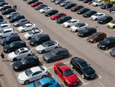 Elevated wide-angle shot of an outdoor car show in a large parking area, rows of vehicles visible from above in natural daylight, attendees as small figures between the cars