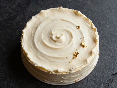 Overhead shot of a minimalist vanilla bean cake with smooth cream frosting, garnished with edible gold leaf on a dark stone surface.