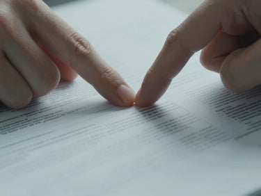 Close-up of a hand pointing at a specific line on a structured printed French document, cool overhead daylight, fingertip resting on the page, crisp focus on the paper texture, no faces visible