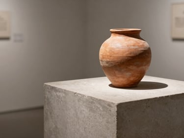 Minimalist gallery installation, close-up of a raw terracotta vessel on a concrete plinth, soft side lighting, sharp shadows.