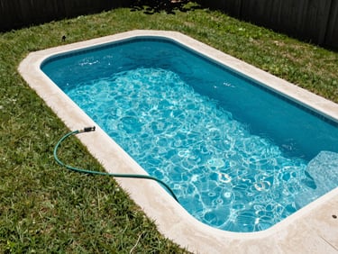 Overhead wide shot of a clear blue backyard pool in a South Shore suburb, bright midday sun, surrounding green lawn, a garden hose coiled neatly beside the pool edge