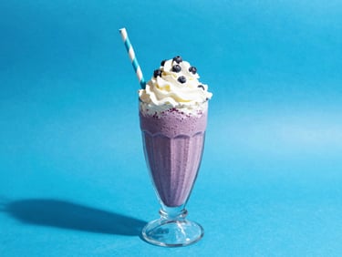 A thick blueberry milkshake in a vintage glass with a striped straw, whipped cream on top, bright blue background.