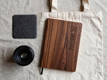 Overhead flat-lay of a curated corporate identity set on a raw linen surface: a dark slate coaster, an engraved walnut notebook, and a heavy-weight canvas tote.