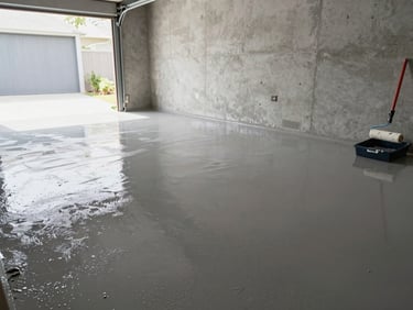 Wide shot of a freshly coated garage floor still wet under natural daylight streaming through an open garage door, roller and coating tray visible to the side, clean concrete walls in background, no people, straightforward documentary framing emphasizing the sealed surface
