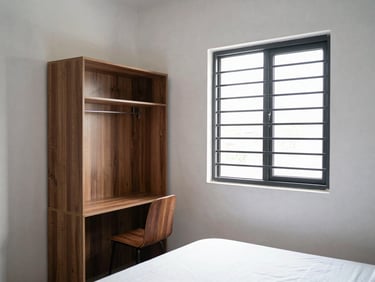 A clean, bright single-occupancy ladies PG room in Yelahanka showing a study corner, a wardrobe, and a secure window with safety grills.