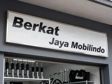 Eye-level crisp photography of Berkat Jaya Mobilindo storefront in Bekasi, clean metallic signage, organized shelves of shock absorbers and tie rods visible inside, bright daylight, industrial texture