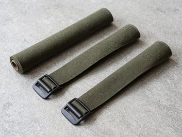 Flat-lay of raw heavy-duty olive green canvas rolls and matte black metal buckles arranged on a clean concrete workspace surface