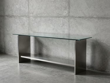 Wide environmental shot of a minimalist furnished room showing a steel and glass console table against a bare concrete wall, cool north-facing daylight, no styling props, the structural legs and shelf clearly visible from a three-quarter angle