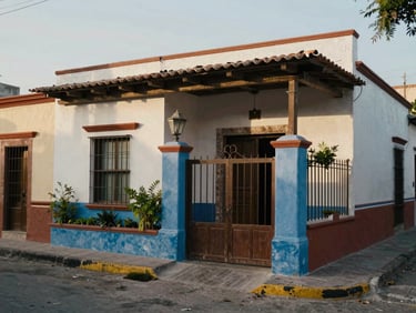 Exterior of a compact house in a quiet Mexican street, afternoon diffused light, painted gate, small garden strip at entry, no people