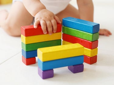 Close-up of toddler hands stacking colorful wooden arches on a clean white floor, warm daylight
