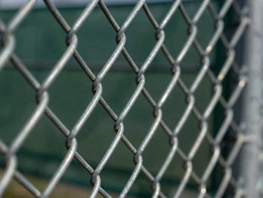 Sharp macro shot of heavy-gauge galvanized chain link fence, showing tightly woven wires and strong tensioning, metallic textures highlighted by harsh sun.