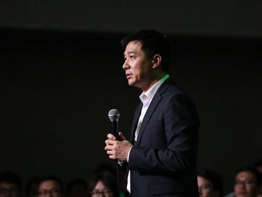 Low-key cinematic shot of a professional male speaker on a dimly lit stage, subtle green ambient spill, shallow depth of field, 35mm