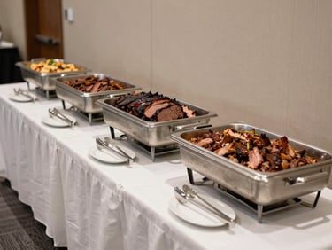 Wide shot of a corporate reception buffet setup indoors — chafing dishes of smoked brisket and pulled pork on a long draped table, tongs resting across trays, professional event lighting, no staff or guests present, clean documentary framing