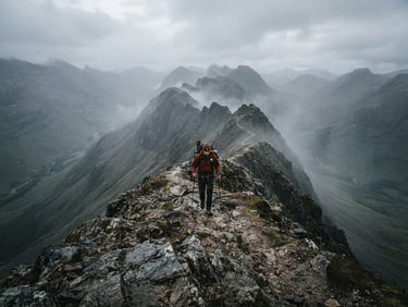 Wide-angle environmental shot of a hiker navigating a narrow mountain ridge under misty morning light, desaturated forest greens and slate grays.