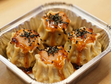 Appetizing close-up of a small foil box containing four pieces of dimsum, thick orange mentai sauce heavily torched on top, sprinkled with nori, warm light