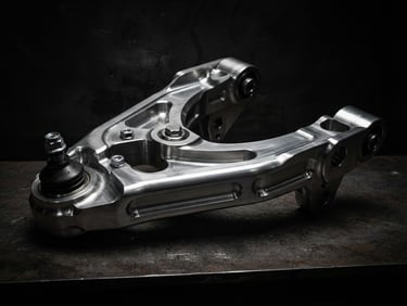 Low-angle high-contrast studio shot of a machined billet steel suspension control arm, sharp metallic highlights, deep shadows, dark background.