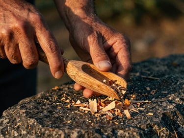 Extreme close-up of weathered hands carving a cedar spoon with a traditional knife, wood shavings falling onto a dark granite rock, warm low golden hour light.